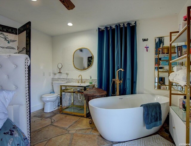 bathroom featuring a freestanding tub, toilet, recessed lighting, a sink, and stone tile flooring