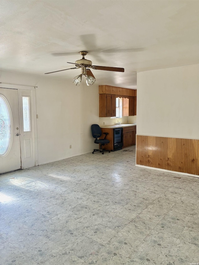 unfurnished living room featuring wooden walls, a ceiling fan, a wainscoted wall, wine cooler, and light floors