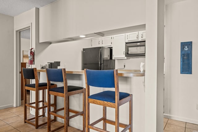 kitchen featuring a breakfast bar area, light tile patterned floors, stainless steel microwave, freestanding refrigerator, and white cabinetry