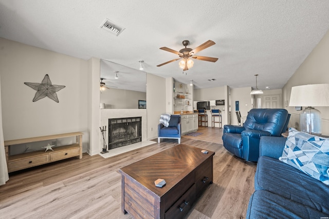 living area with a textured ceiling, ceiling fan, a fireplace, visible vents, and light wood finished floors