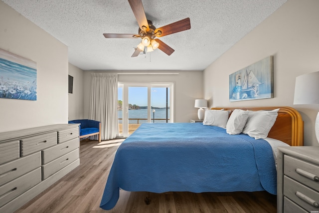 bedroom with a ceiling fan, wood finished floors, a water view, access to exterior, and a textured ceiling