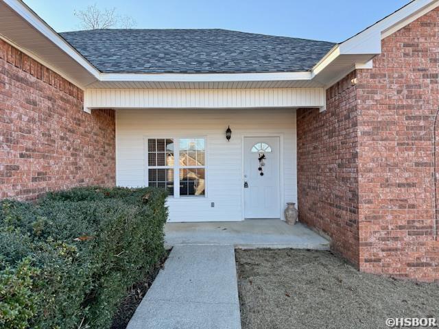 view of exterior entry featuring brick siding, a patio, and roof with shingles