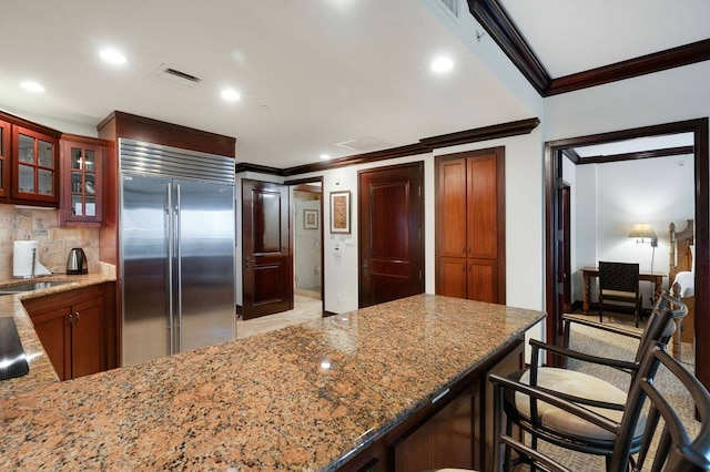 kitchen featuring light stone counters, ornamental molding, backsplash, a kitchen bar, and stainless steel built in fridge