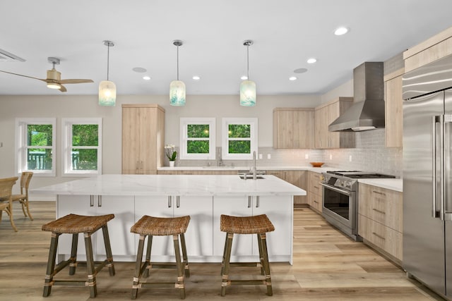 kitchen with high quality appliances, wall chimney exhaust hood, decorative backsplash, light brown cabinetry, and light stone counters