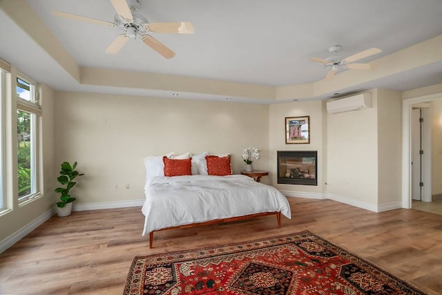 bedroom featuring a raised ceiling, a wall mounted air conditioner, ceiling fan, and light wood-type flooring