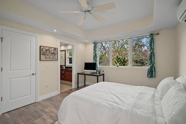 bedroom with light wood-type flooring, an AC wall unit, ceiling fan, connected bathroom, and a raised ceiling