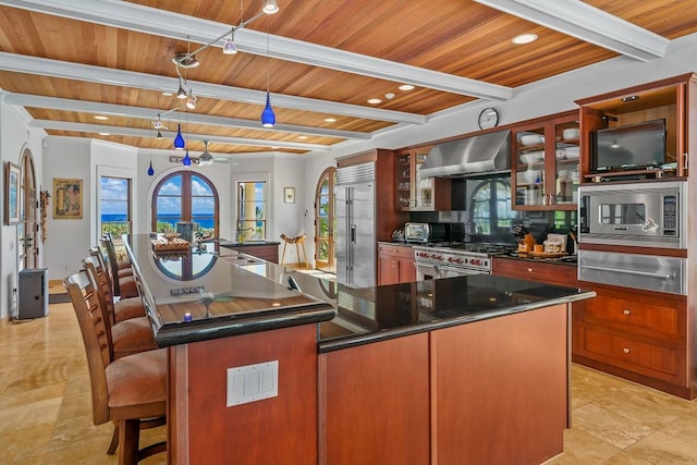 kitchen with built in appliances, beamed ceiling, a center island with sink, wooden ceiling, and wall chimney range hood