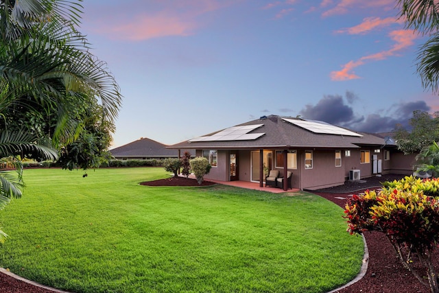 back house at dusk with a patio area, central AC unit, solar panels, and a yard