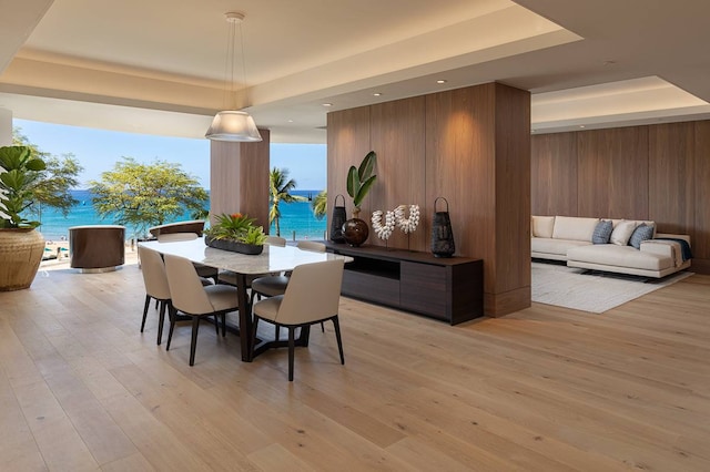 dining room with wood walls, a water view, a tray ceiling, and light hardwood / wood-style floors