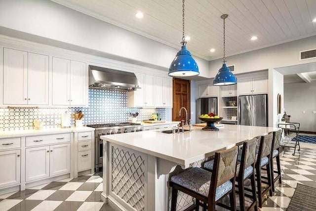 kitchen featuring an island with sink, light tile floors, appliances with stainless steel finishes, wall chimney exhaust hood, and white cabinetry