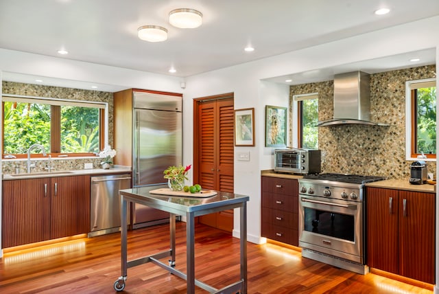 kitchen featuring wall chimney range hood, high quality appliances, hardwood / wood-style floors, sink, and tasteful backsplash