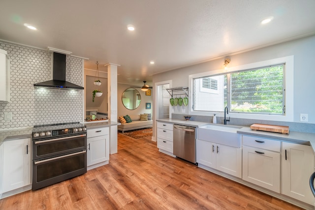 kitchen featuring wall chimney range hood, light hardwood / wood-style flooring, backsplash, stainless steel dishwasher, and double oven range