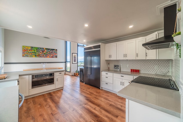 kitchen with oven, backsplash, wall chimney exhaust hood, white cabinets, and built in refrigerator