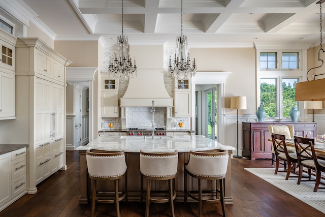 kitchen with premium range hood, coffered ceiling, a center island with sink, backsplash, and dark wood-type flooring