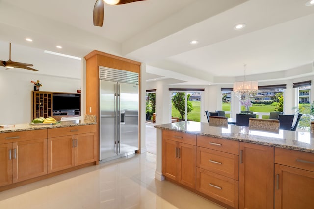 kitchen featuring light stone countertops, stainless steel built in fridge, hanging light fixtures, and ceiling fan with notable chandelier