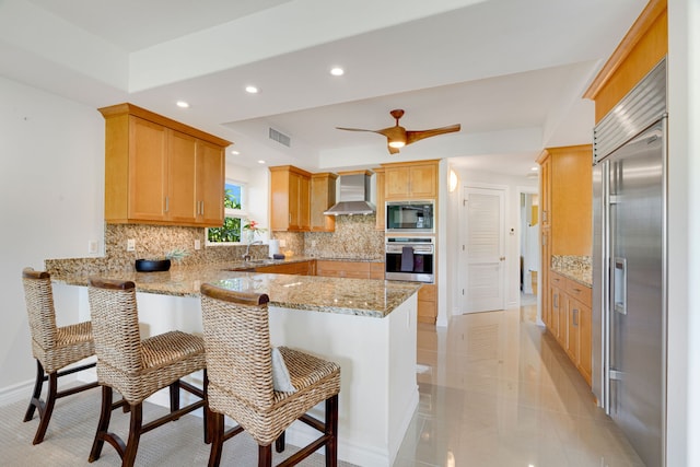 kitchen featuring wall chimney range hood, tasteful backsplash, light stone counters, kitchen peninsula, and built in appliances