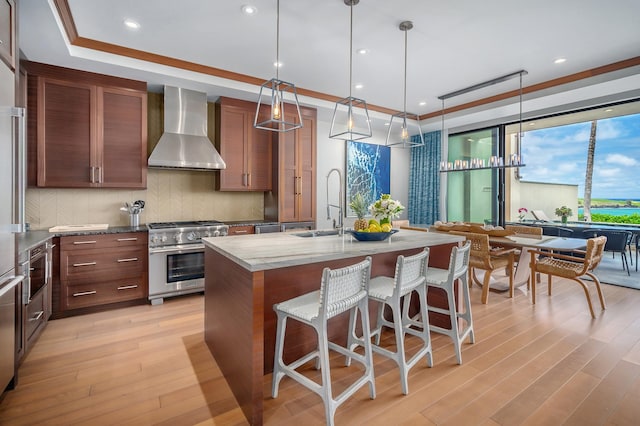 kitchen with stainless steel stove, a raised ceiling, a center island with sink, and wall chimney range hood