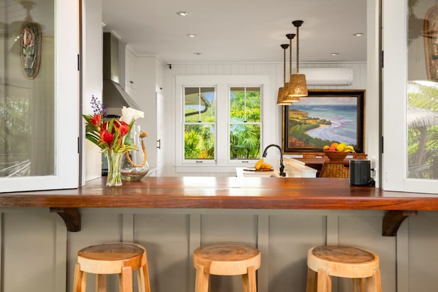 bar featuring wall chimney range hood, sink, a wall unit AC, white cabinets, and decorative light fixtures