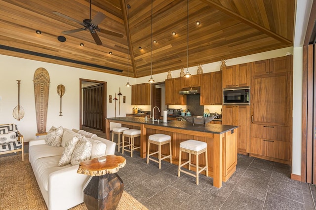 kitchen with black microwave, a breakfast bar area, under cabinet range hood, brown cabinetry, and dark countertops