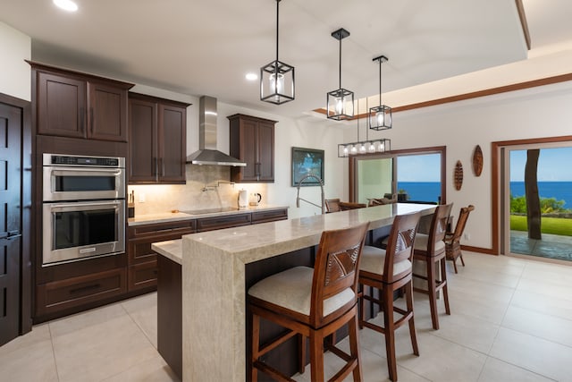 kitchen with a water view, wall chimney exhaust hood, an island with sink, double oven, and a breakfast bar