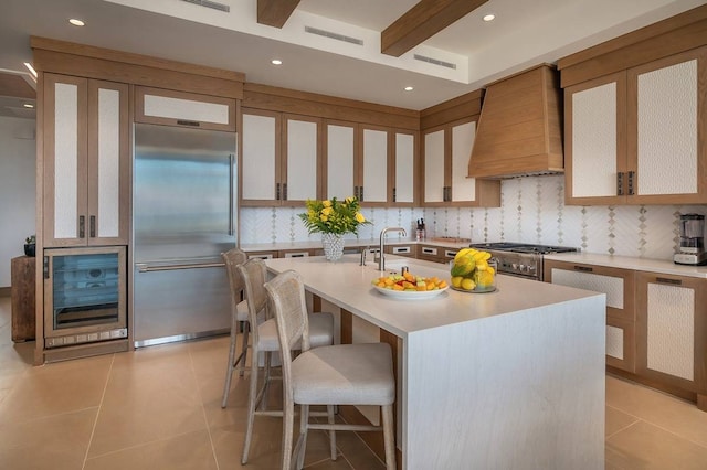 kitchen featuring premium appliances, beverage cooler, a center island with sink, custom range hood, and decorative backsplash