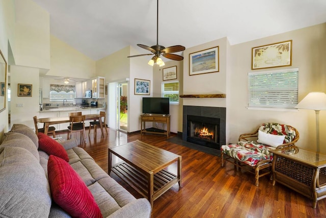 living room with high vaulted ceiling, sink, dark wood-type flooring, a tiled fireplace, and ceiling fan