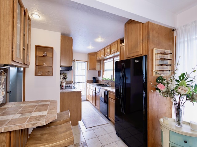 kitchen with light tile patterned floors, tile countertops, glass insert cabinets, black appliances, and a sink