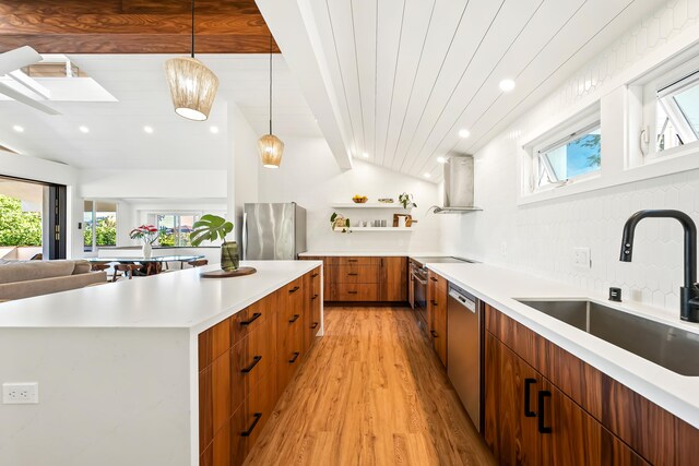 kitchen featuring sink, light wood-type flooring, appliances with stainless steel finishes, wall chimney exhaust hood, and hanging light fixtures