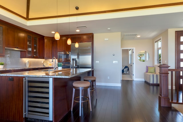 kitchen with an island with sink, hanging light fixtures, beverage cooler, built in appliances, and light stone countertops