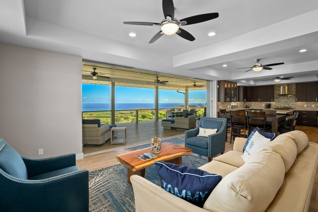 living room with a raised ceiling, light hardwood / wood-style flooring, ceiling fan, and a water view