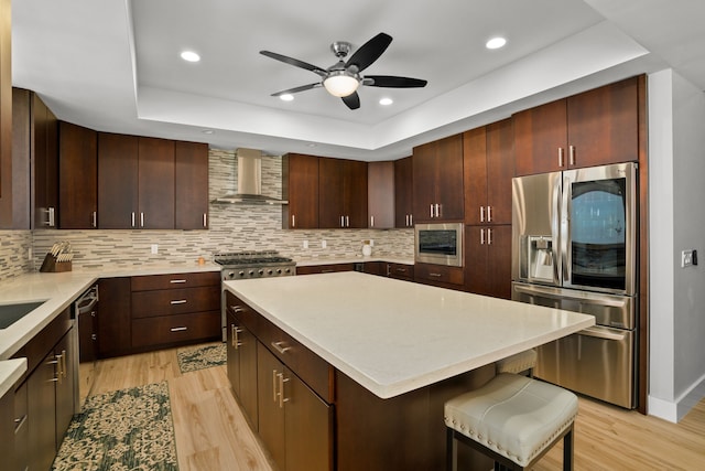 kitchen featuring light wood-type flooring, dark brown cabinets, ceiling fan, appliances with stainless steel finishes, and wall chimney range hood