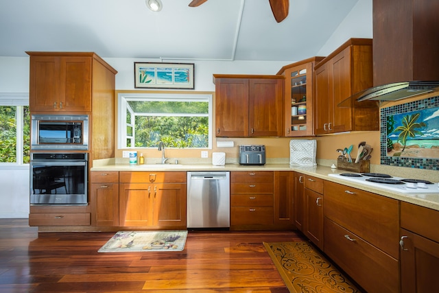 kitchen with stainless steel appliances, brown cabinetry, vaulted ceiling, wall chimney range hood, and light countertops