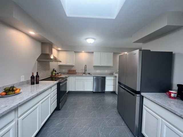 kitchen with white cabinetry, light stone counters, stainless steel appliances, sink, and wall chimney range hood