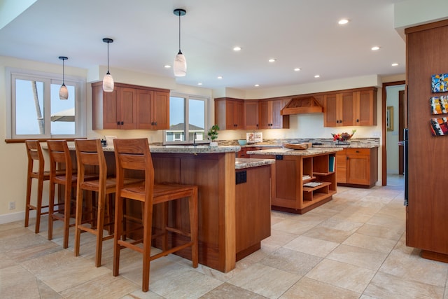 kitchen with pendant lighting, a kitchen island, light stone counters, a kitchen bar, and wall chimney exhaust hood