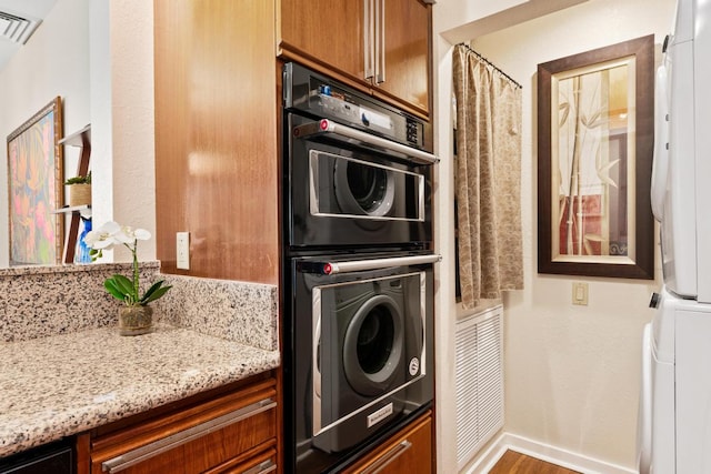 kitchen with stacked washer / drying machine, black double oven, hardwood / wood-style flooring, and light stone countertops