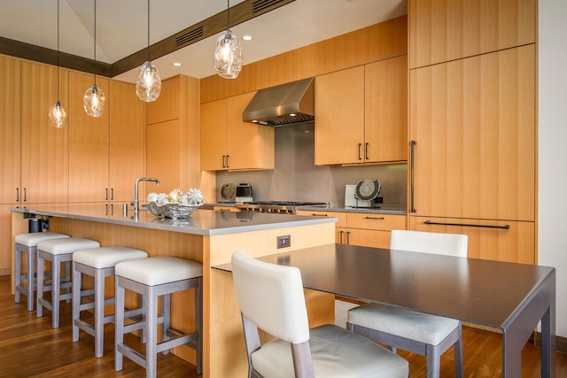 kitchen with light brown cabinets, a center island with sink, dark wood-type flooring, and range hood