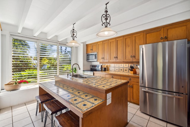 kitchen with a kitchen island with sink, beamed ceiling, stainless steel appliances, light tile patterned flooring, and decorative light fixtures