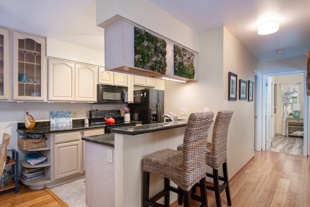 kitchen with kitchen peninsula, light wood-type flooring, dark stone counters, black appliances, and a breakfast bar area