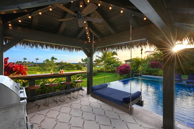 pool at dusk featuring a lawn, ceiling fan, grilling area, a gazebo, and a patio area