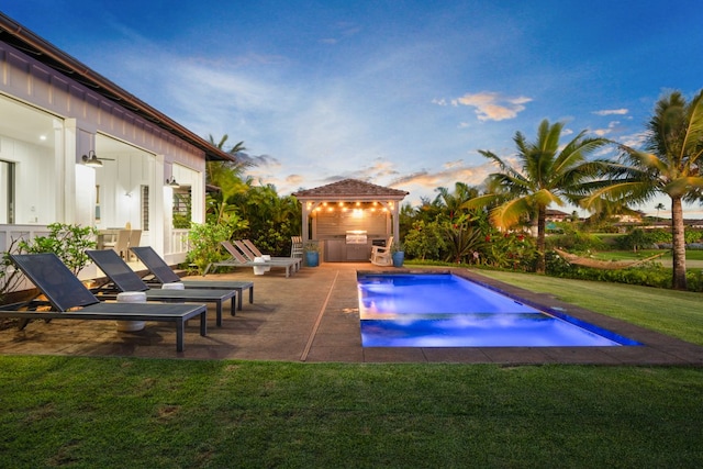 pool at dusk with a lawn, an outdoor kitchen, ceiling fan, and a patio area