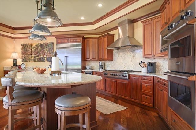 kitchen with dark hardwood / wood-style flooring, ornamental molding, wall chimney exhaust hood, stainless steel appliances, and an island with sink