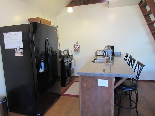 kitchen with sink, a kitchen breakfast bar, stainless steel gas range oven, black refrigerator with ice dispenser, and hardwood / wood-style floors