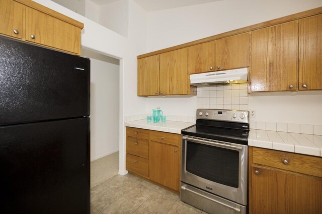 kitchen with electric range, under cabinet range hood, freestanding refrigerator, brown cabinetry, and tile counters