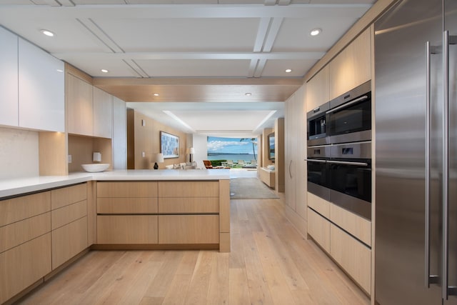 kitchen with light brown cabinets, coffered ceiling, built in fridge, light wood-type flooring, and kitchen peninsula