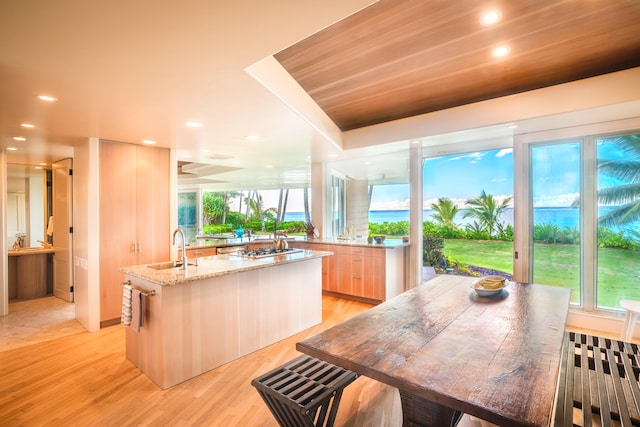 kitchen with light wood-type flooring, light stone countertops, a kitchen island with sink, and light brown cabinetry