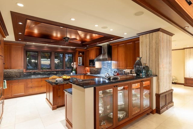 kitchen featuring backsplash, dark stone counters, wall chimney exhaust hood, a raised ceiling, and a kitchen island