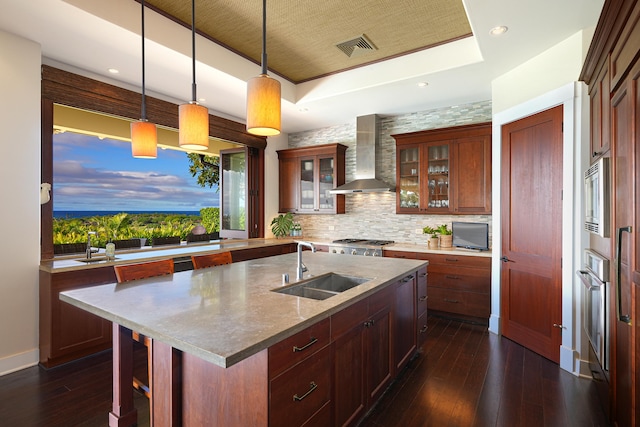 kitchen with sink, hanging light fixtures, wall chimney range hood, dark hardwood / wood-style flooring, and a kitchen island with sink