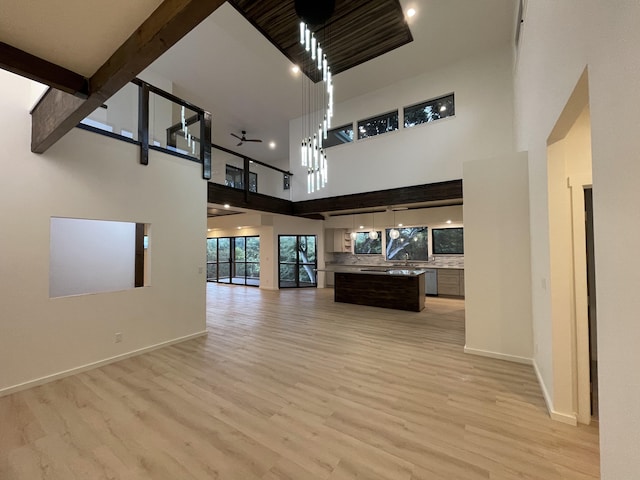 unfurnished living room featuring beam ceiling, ceiling fan, a towering ceiling, and light hardwood / wood-style floors