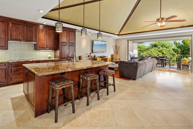 kitchen featuring a kitchen bar, hanging light fixtures, an island with sink, light stone countertops, and decorative backsplash