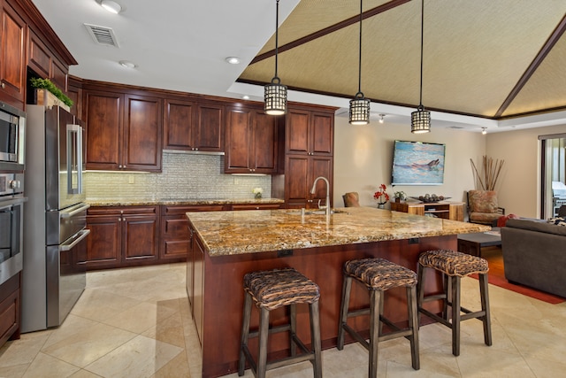 kitchen featuring sink, hanging light fixtures, stainless steel appliances, light stone counters, and an island with sink